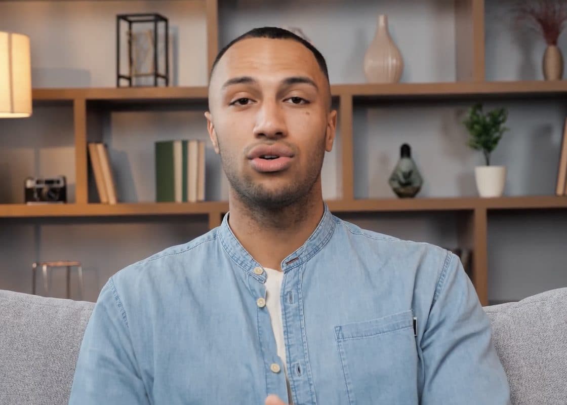 A man on a sofa in front of a bookshelf, talking to the camera.