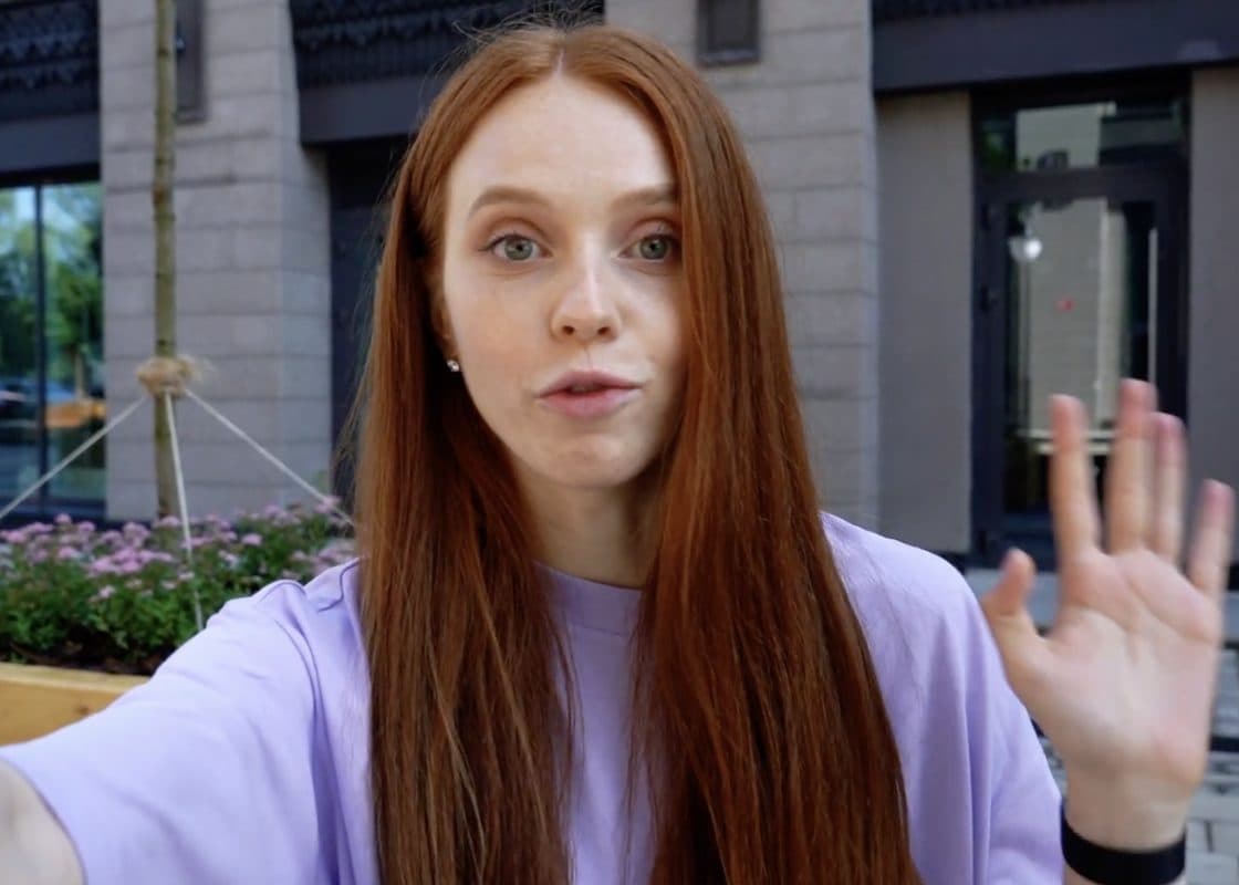 A woman outdoors waving hello to her phone camera.